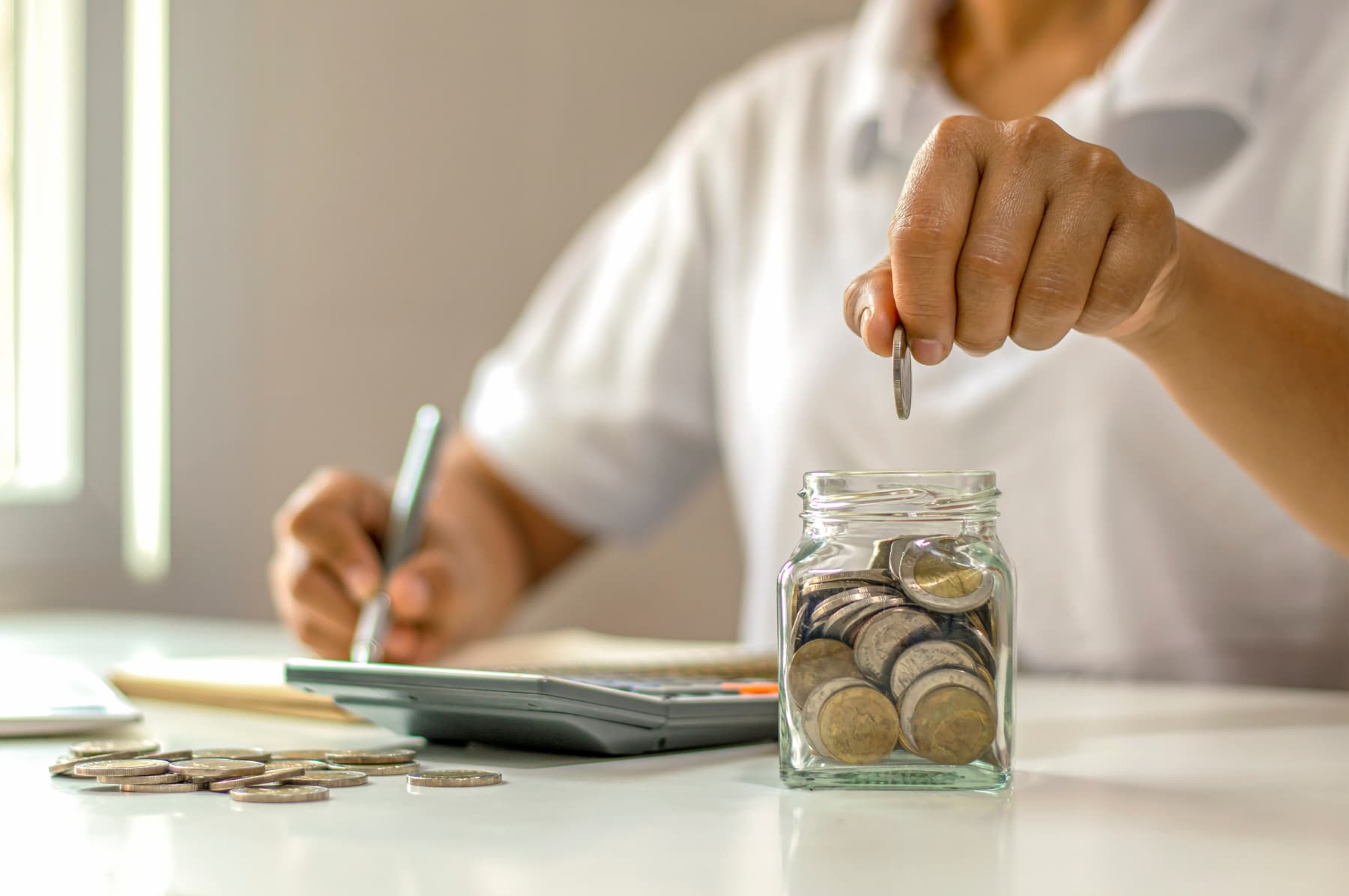 Persona escribiendo notas en un cuaderno financiero, con una calculadora en la mesa y metiendo una moneda en un frasco, simbolizando la importancia de ahorrar y minimizar gastos innecesarios para una mejor automatización y eficiencia en el control financiero.