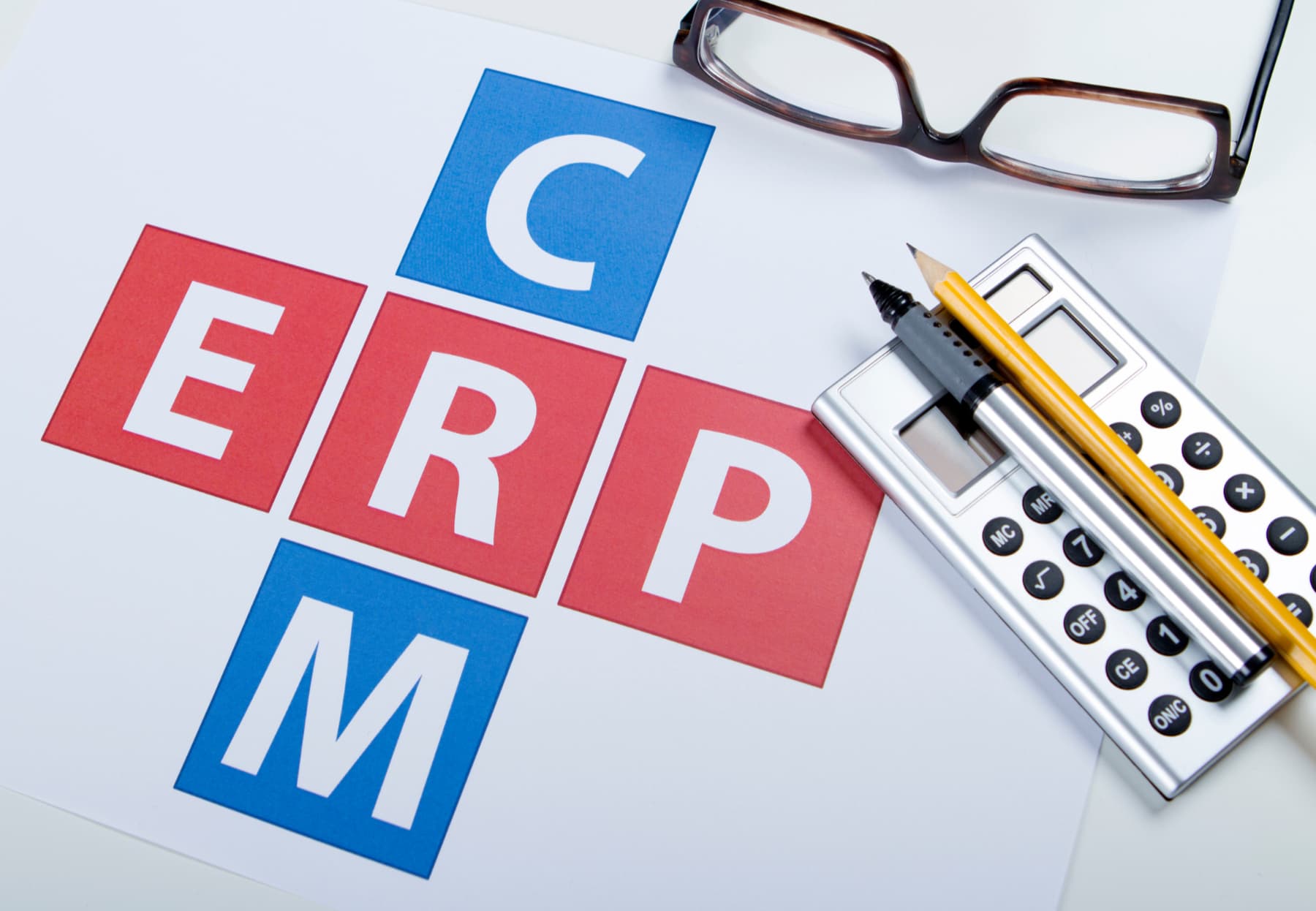 Crucigrama con las palabras 'CRM' y 'ERP' intersectadas, acompañado de unos lentes y un lápiz, simbolizando la importancia de estos sistemas para la gestión empresarial