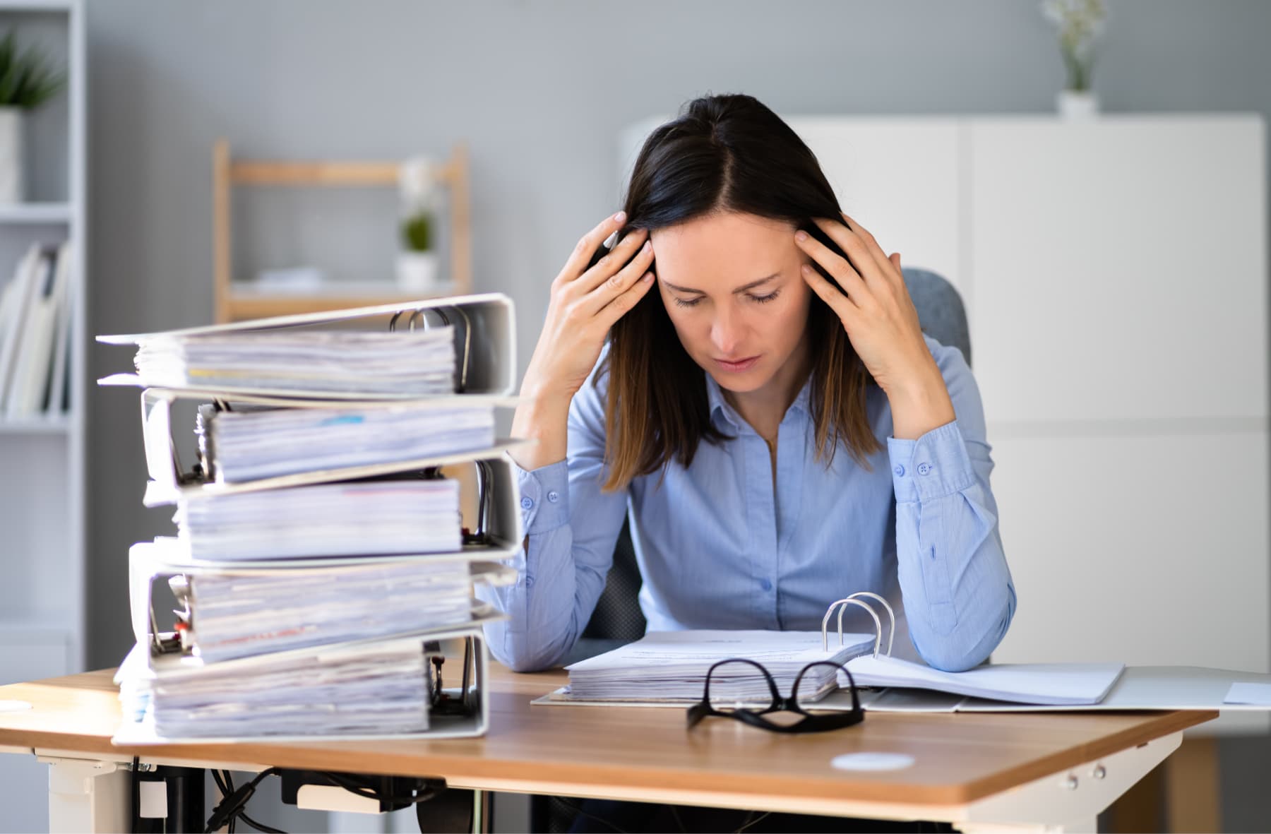 Mujer frustrada con dolor de cabeza, sentada junto a una pila de libros contables, simbolizando el estrés de la contabilidad manual.