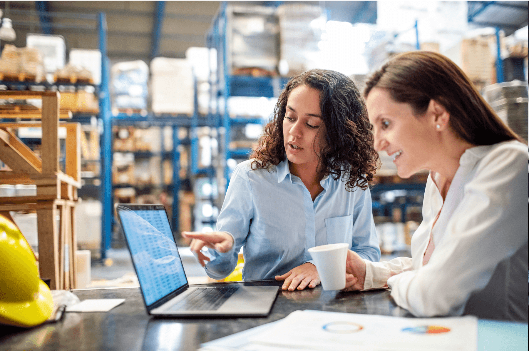 Dos mujeres en un almacén revisando su laptop con gráficas cerca, contentas de que su sistema ERP les permita tener un control integral sobre sus entradas y salidas.
