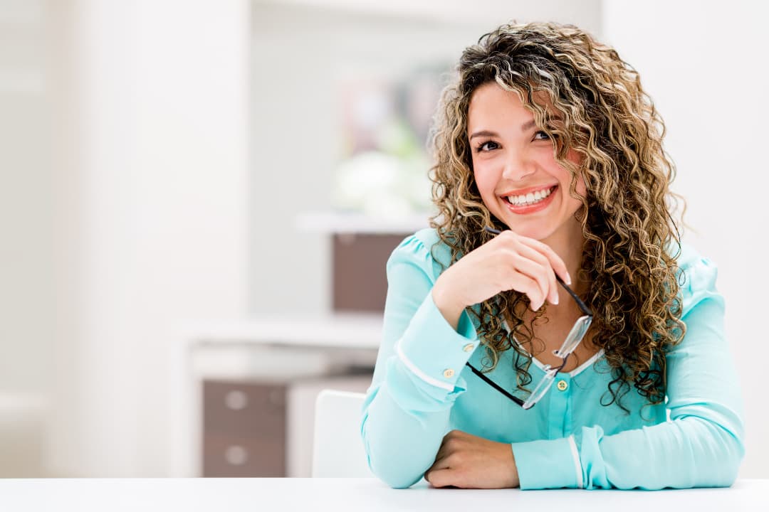 Mujer de negocios con blusa celeste y cabello rizado, sosteniendo sus lentes y sonriendo con satisfacción, reflejando la felicidad por el florecimiento de su negocio gracias a la organización que le permite su sistema ERP.
